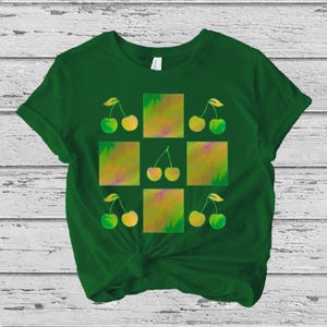 以下が含まれることがあります： グリッド状にチェリーのパターンが施された緑色のTシャツ。チェリーは緑と黄色で、抽象的なカラフルな正方形の背景に配置されています。