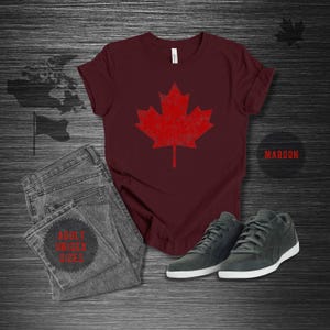 K&ouml;nnte beinhalten: Burgunderfarbenes T-Shirt mit einem roten Ahornblatt-Design im Used-Look. Das Bild zeigt graue Jeans und dunkelgraue Turnschuhe. Der Text "ADULT UNISEX SIZES" ist auf der Jeans zu sehen. Das Wort "MAROON" ist ebenfalls sichtbar.