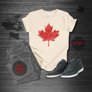 K&ouml;nnte beinhalten: Cremefarbenes T-Shirt mit einem roten Ahornblatt-Grafik-Design, kombiniert mit grauen Jeans und dunkelgrauen Turnschuhen. Das Shirt ist mit "NATURAL" und "ADULT UNISEX SIZES" beschriftet.