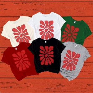 May include: Six t-shirts in different colors with a red flower design made of the words "Merry Christmas". The shirts are laid out on a red wooden background.