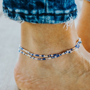May include: A beaded anklet with blue and white beads. The anklet is worn on an ankle, with blue denim jeans visible in the background. The anklet is made of small, round beads strung together.