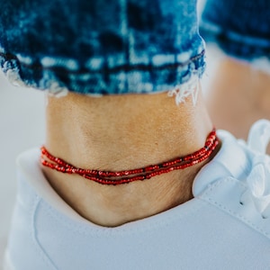 May include: A close-up of a red beaded ankle bracelet. The bracelet is worn on an ankle, with a white sneaker in the foreground and blue jeans in the background. The bracelet has a delicate, layered design.