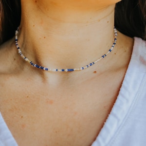 Op de afbeelding: Een delicate kralenketting met kleine blauwe, witte en crèmekleurige kralen. De ketting wordt om de nek gedragen, tegen een wit overhemd. De kralen zijn gelijkmatig verdeeld, wat een subtiel, elegant ontwerp creëert.