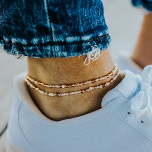 May include: Close-up of three delicate beaded anklets in gold, white, and copper tones, worn on an ankle above a white sneaker. The anklets feature small beads strung together, creating a layered effect. The person is wearing blue denim jeans.