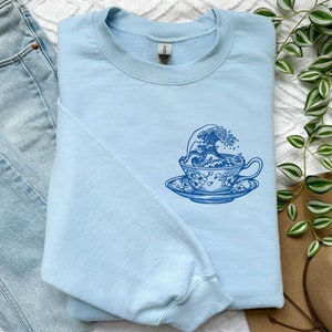 Könnte beinhalten: Ein hellblaues Sweatshirt mit einer blauen Grafik einer Welle, die in eine Teetasse kracht. Die Teetasse hat ein florales Design.