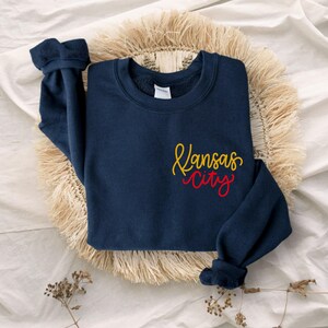 Puede incluir: Una sudadera azul marino con las palabras "Kansas City" en letras amarillas y rojas.