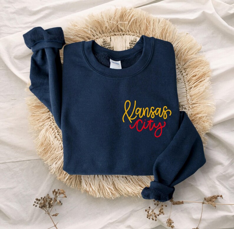 Puede incluir: Una sudadera azul marino con las palabras "Kansas City" en letras amarillas y rojas.