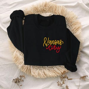 Puede incluir: Sudadera negra con un dise&ntilde;o bordado en dorado y rojo que dice "Kansas City".