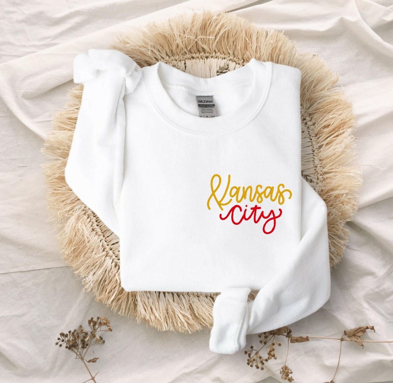 Puede incluir: Sudadera blanca con un gr&aacute;fico de "Kansas City" en dorado y rojo.