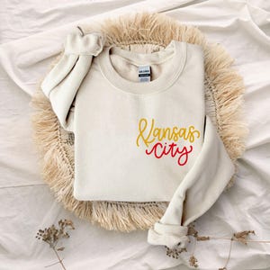 Puede incluir: Una sudadera de color crema con las palabras "Kansas City" escritas en rojo y amarillo.