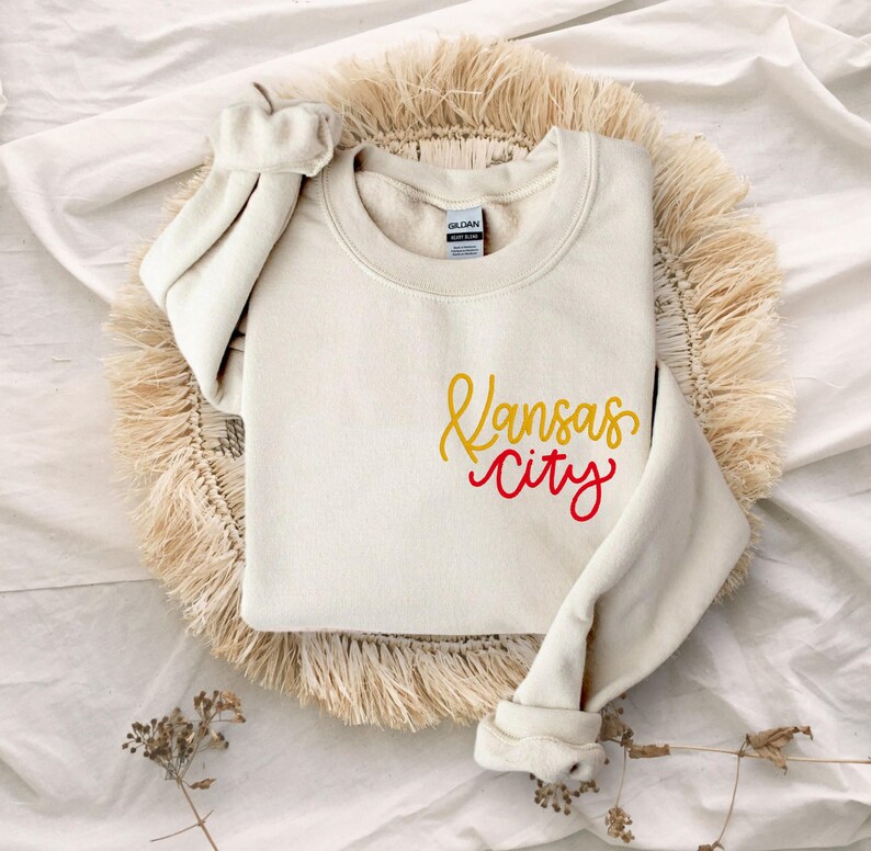 Puede incluir: Una sudadera de color crema con las palabras "Kansas City" escritas en rojo y amarillo.