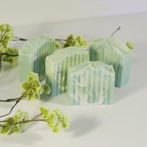 May include: Four bars of handmade soap with a green and white swirl design. The soap is arranged on a white surface with green sprigs of foliage.