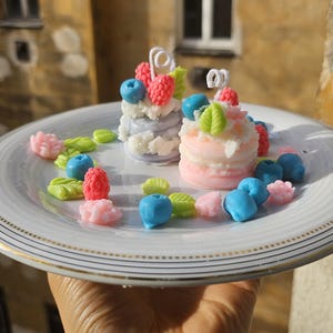 Puede incluir: Dos pasteles en miniatura decorados con glaseado rosa, azul y verde y cubiertos con arándanos y frambuesas. Los pasteles están en un plato blanco con un borde dorado, rodeados de pequeñas flores y bayas decorativas.