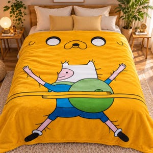 May include: A bright yellow plush blanket featuring a large graphic print of the character Jake the Dog's face at the top and a full-body illustration of the character Finn the Human holding a green circular object in the center.