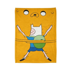 May include: A yellow fleece blanket featuring a cartoon image of Finn the Human from Adventure Time being swallowed by a giant yellow dog. Finn is wearing a blue shirt and a green backpack.