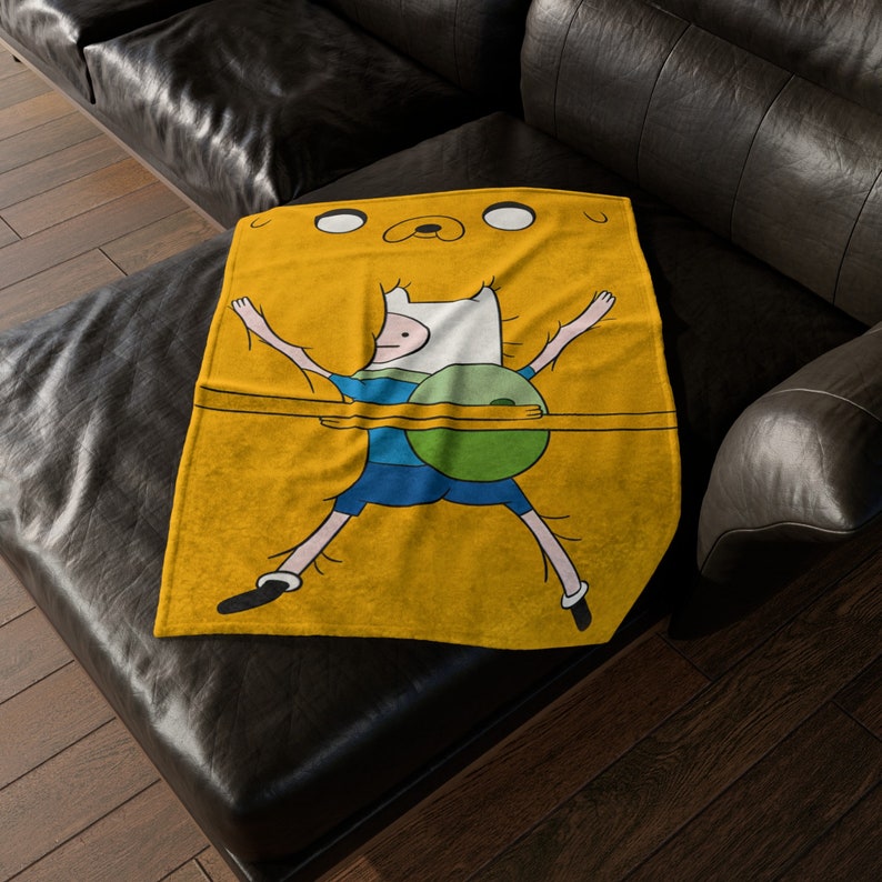 May include: A yellow fleece throw blanket featuring a cartoon image of Finn the Human from Adventure Time. Finn is wearing a blue shirt and green trousers and is stretched out with his arms and legs extended.