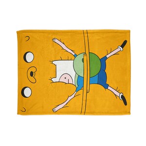 May include: A yellow fleece blanket featuring a cartoon character from Adventure Time, Finn the Human. Finn is lying on his back with his arms and legs outstretched.