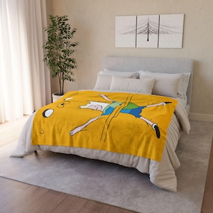 May include: A yellow fleece throw blanket featuring a cartoon image of Finn the Human from Adventure Time. Finn is lying on his back with his arms and legs outstretched.