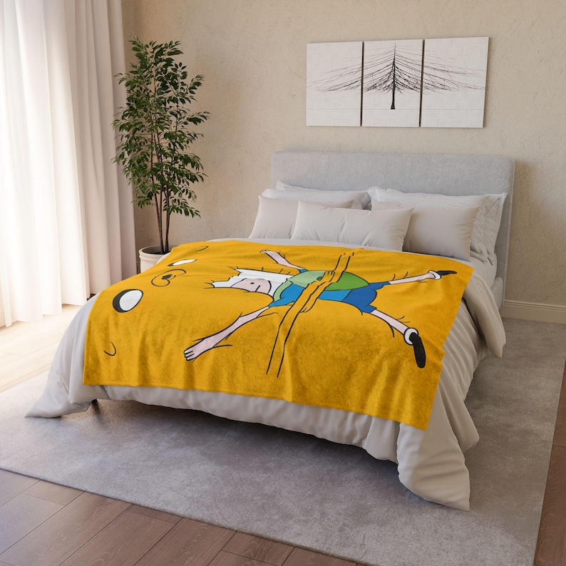 May include: A yellow fleece throw blanket featuring a cartoon image of Finn the Human from Adventure Time. Finn is lying on his back with his arms and legs outstretched.