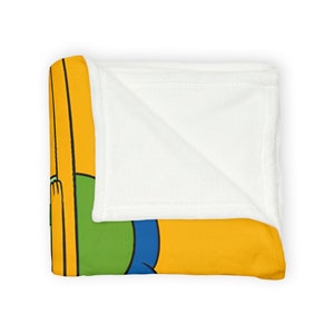 May include: A fleece blanket featuring a cartoon illustration of a person in a yellow, green, and blue outfit. The blanket is folded in half, revealing the white fleece lining.