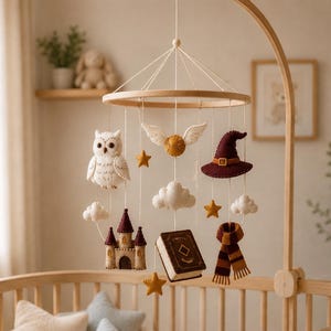 May include: A hanging nursery mobile featuring felted figures suspended from a circular wooden frame. The figures include a white owl, a winged golden ball, a maroon witch hat, a castle, a brown book, a striped scarf, white clouds, and stars.