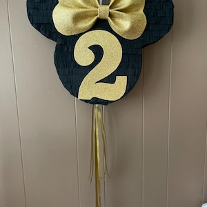 May include: Black and gold piñata in the shape of a mouse head with a large bow and the number 2. The piñata has gold streamers hanging from the bottom. The background is a light brown wall.