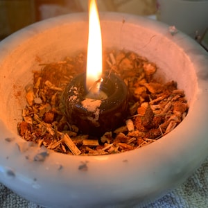 Può includere: Una ciotola in ceramica bianca riempita con pezzi botanici essiccati e trucioli di legno, con un portacandele centrale marrone scuro contenente una candela accesa. La fiamma arancione e gialla proietta una luce calda sul contenuto.