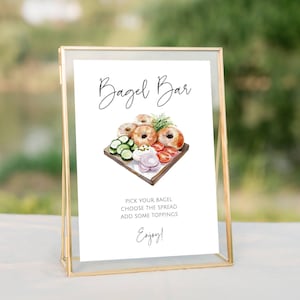 May include: A gold-framed sign with the words "Bagel Bar" in elegant script. The sign features a watercolor illustration of bagels, sliced cucumbers, tomatoes, and toppings. The text below reads "Pick your bagel, choose the spread, add some toppings, enjoy!"