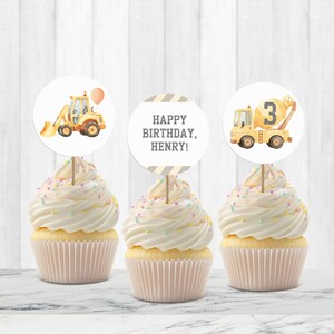 May include: Three cupcakes with white frosting and colorful sprinkles. Each cupcake has a round topper with a construction theme. One topper shows a yellow excavator, another says "HAPPY BIRTHDAY, HENRY!", and the third shows a cement mixer with the number 3.