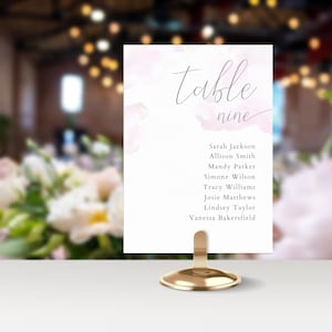 May include: A rectangular white table seating card featuring soft pink watercolor accents and the text Table nine, followed by a list of names including Sarah Jackson, Allison Smith, Mandy Parker, Simone Wilson, Tracy Williams, and others, held by a gold stand.