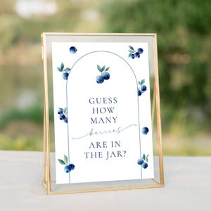 May include: A framed sign with the text "Guess How Many Berries Are In The Jar?" in navy blue. The sign is decorated with watercolor blueberries and green leaves. The frame is gold.