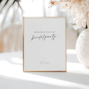 May include: A framed sign with gold trim and white background. The sign reads "RESERVED FOR THE bridal party" in elegant script. Below the text are three dots, followed by names and a date. The sign is a wedding or event decoration.