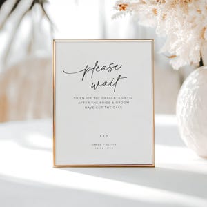 May include: A framed sign with the words "please wait" in elegant script. The text below reads "TO ENJOY THE DESSERTS UNTIL AFTER THE BRIDE & GROOM HAVE CUT THE CAKE." The bottom of the sign includes names and a date. The frame is gold.