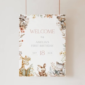 May include: A welcome sign for Amelia's first birthday, featuring illustrations of a raccoon, bear, giraffe, bunny, and cat reading books. The sign includes the date: August 18th.