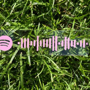 May include: A clear acrylic Spotify code tag with a pink Spotify logo and pink soundwave pattern. The tag is rectangular and rests on a bed of green grass.