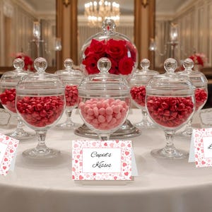 Op de afbeelding: Een tafelsetting met glazen potten gevuld met rode hartvormige snoepjes en roze snoepjes. Een grote glazen kom bevat rode rozen. Kleine kaarten met tekst liggen op de tafel.
