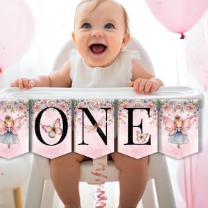 May include: A banner with the word "ONE" is displayed in front of a baby in a high chair. The banner has a pink background with floral and fairy illustrations. Pink balloons are in the background, creating a festive atmosphere.