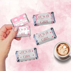 May include: Pink and silver wrapped candies with "IT'S A GIRL" printed on them. One candy has the text "Welcoming our new bundle of joy!". A cup of coffee with latte art is in the background.