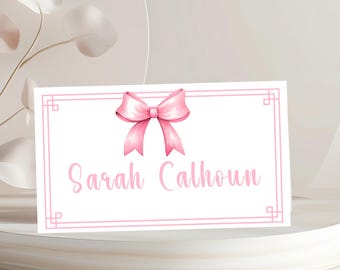 May include: A rectangular white card featuring a pink double-lined border with geometric corner accents. A pink ribbon bow is centered at the top, and the name Sarah Calhoun is printed in a pink cursive script font in the center.