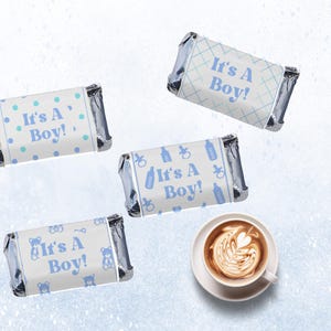 Könnte beinhalten: Vier Mini-Schokoriegel mit blau-weißen Verpackungen, jeweils mit dem Text "It's a Boy!". Designs umfassen Punkte, ein Gittermuster, Babyflaschen und Teddybären. Eine Tasse Kaffee mit Latte Art ist ebenfalls vorhanden.