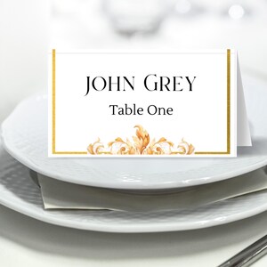 May include: White place card with a gold border and a gold decorative design at the bottom. The text on the card reads "JOHN GREY Table One".