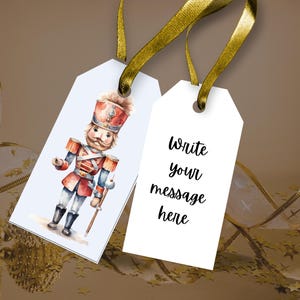 May include: Two white rectangular gift tags with gold ribbon loops. One tag features a watercolor illustration of a nutcracker soldier in a red and blue uniform. The other tag has the black printed text "Write your message here" in the center.