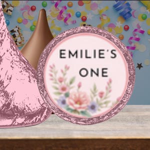 May include: A pink and brown Hershey's Kiss candy next to a round pink label with the text "EMILIE'S ONE" and floral accents. The background features confetti and streamers, suggesting a birthday celebration.