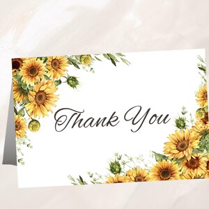 May include: A white folded card with a floral design featuring yellow sunflowers and green leaves. The words "Thank You" are written in elegant black script across the center of the card. The sunflowers are arranged around the edges.