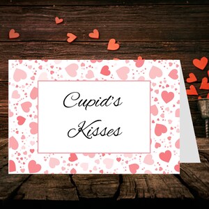 Op de afbeelding: Een Valentijnsdagkaart met een patroon van roze harten en de tekst "Cupid's Kisses". De kaart ligt op een houten oppervlak, met hartvormige decoraties verspreid.