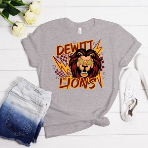 May include: A heather grey t-shirt featuring a graphic design of a lion wearing star-shaped sunglasses. The graphic also includes the text "DEWITT LIONS" in yellow and red.