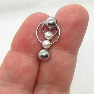 Puede incluir: Una pieza de joyería plateada para piercing corporal con dos pequeñas perlas blancas y dos bolas plateadas. La joya tiene dos anillos plateados y está sujeta por el dedo de una persona.
