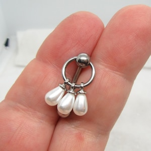 Könnte beinhalten: Ein silberfarbenes Barbell-Piercing mit einem hängenden Design aus drei weißen, birnenförmigen Kunstperlen. Das Barbell hat einen kreisförmigen Ring und eine kleine Kugel an der Oberseite.