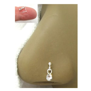 May include: A silver nose ring with a dangling clear crystal. The nose ring is shown on a person's nose. A smaller inset image shows the nose ring held by a finger.