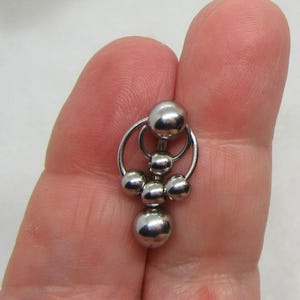 May include: A silver-colored body piercing jewelry piece. The jewelry features a barbell design with multiple spherical beads and circular accents. The piece is held in a hand against a white background.